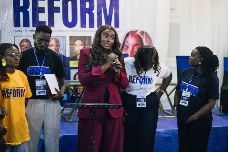 The speaker in a maroon suit addressing the room with conference staff beside her